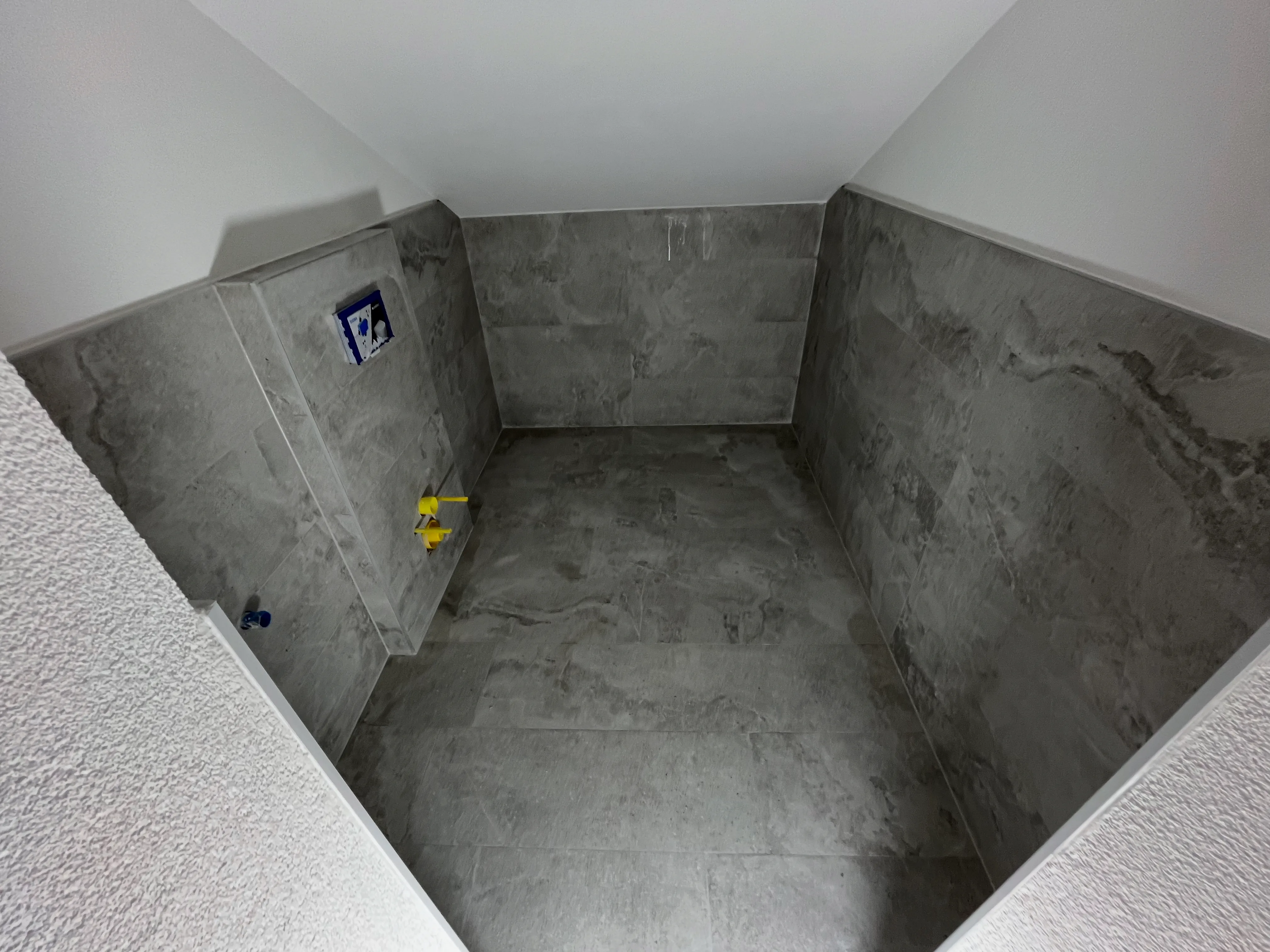 Raum mit grauen Wand- und Bodenfliesen, Vorbereitungen für Sanitärinstallationen sichtbar