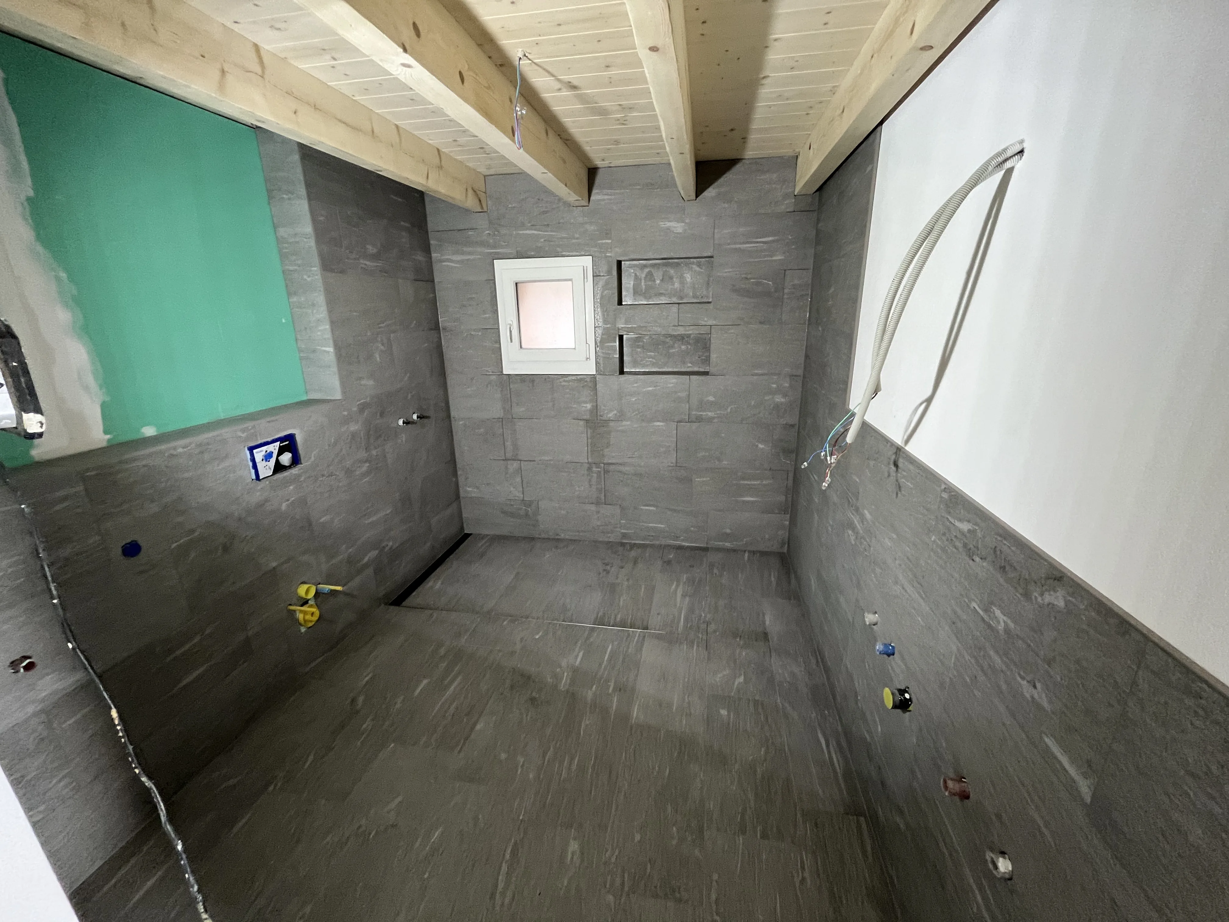 Badezimmer mit grauen Wandplatten, Holzdecke und Fenster, vorbereitet für Fliesenarbeiten